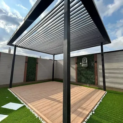 This project showcases a modern, minimalist pergola with a black metal frame and a slatted roof for partial shade. It is installed over a newly laid composite wood deck, framed by artificial turf and a clean white pebble border. This structure is the foundation for a stylish and functional outdoor lounge area.