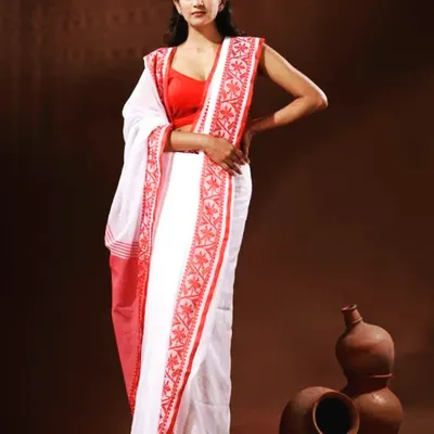 A model wearing the classic white saree with a red embroidered border, paired with a simple red sleeveless blouse. This is a timeless and elegant outfit for any Durga Puja celebration.