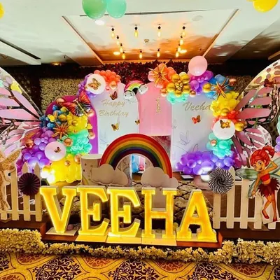 A vibrant butterfly-themed first birthday party for Veeha, featuring a colorful backdrop with large butterfly wings, rainbow accents, and light-up letters.