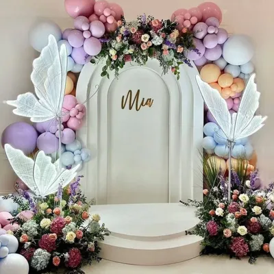 An ethereal and elegant butterfly-themed backdrop for "Mia." This design features a white arched stage, illuminated butterfly wings, and a beautiful arrangement of pastel balloons and fresh flowers, creating a dreamy photo opportunity.
