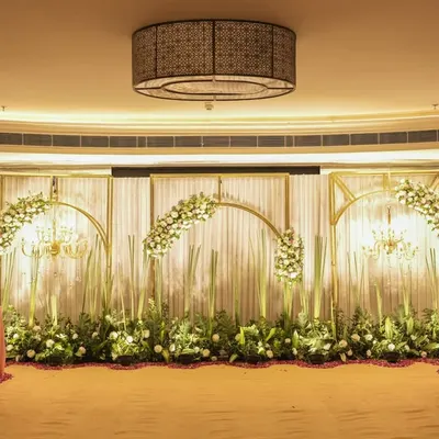 A full view of an elegant indoor stage setup, featuring golden arches, white drapes, and beautiful floral arrangements. This design creates a sophisticated and grand backdrop for a reception or engagement ceremony.