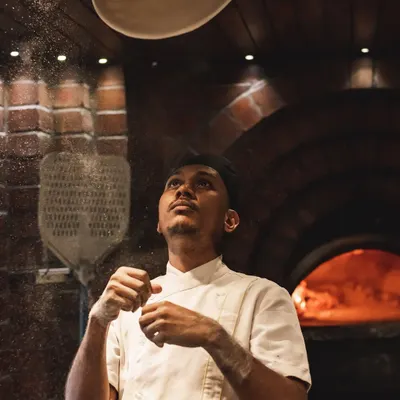A chef expertly tosses a round of pizza dough, with a shower of flour catching the light. The brick oven glows in the background, ready for action.