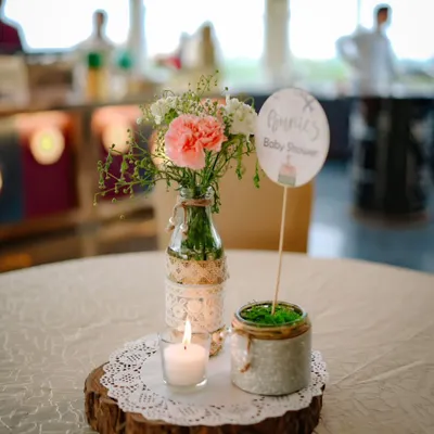 A rustic and charming table centerpiece for a baby shower. A simple arrangement of pink carnations in a lace-wrapped bottle sits on a wood slice next to a small candle, creating a warm, cozy feel.