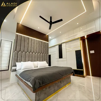 A luxurious master bedroom featuring a floor-to-ceiling grey upholstered headboard with geometric patterns. The design is enhanced by gold trim details on the bed and wardrobe, creating a cohesive and opulent look.