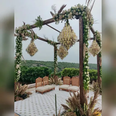 A modern rustic mandap that blends nature with luxury. This outdoor wedding setup features a wooden structure adorned with white roses and greenery, with elegant crystal chandeliers hanging above, all set against a beautiful natural backdrop.
