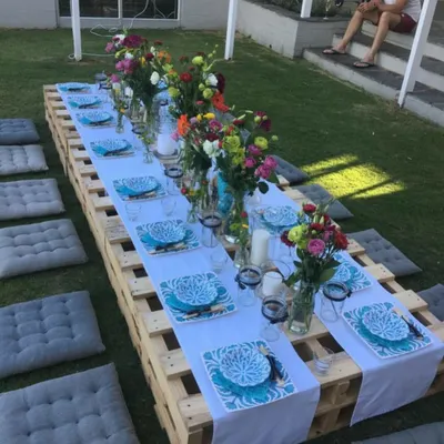 An outdoor picnic style party setup on a lush green lawn. A long, low table made from wooden pallets is set with a white runner, colorful floral arrangements, and bright blue patterned plates, with grey cushions for seating.