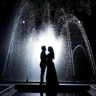 A stunning silhouette of a couple standing before a brightly lit water fountain at night. Their forms are perfectly outlined against the spray, creating a dramatic and romantic image.