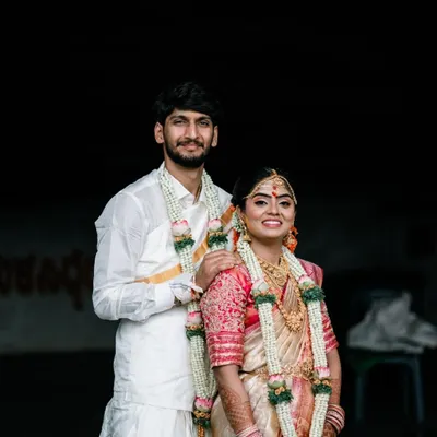 A classic portrait of a South Indian couple on their wedding day. Their traditional white and gold attire, along with the floral garlands, looks beautiful against the simple, dark background.