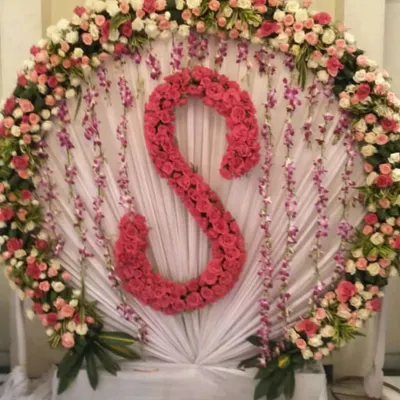 Personalized decorations make any event feel special. This backdrop features a large circular frame of white and pink roses, with the initial 'S' made entirely of bright pink roses as the centerpiece. It's a great idea for birthdays, anniversaries, or engagement parties.