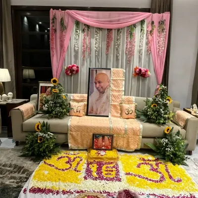 An elegant and serene setup for a Guruji satsang. The decor features a beautiful floral rangoli, sunflower arrangements, and a pink drape backdrop with hanging wisteria to create a divine ambiance.