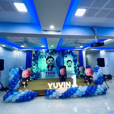 A complete Boss Baby themed stage setup for a first birthday. This includes personalized backdrops with the child's photo, multiple Boss Baby character cutouts, and a blue and white balloon arch.