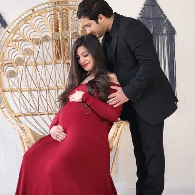 A romantic studio portrait of a couple with a peacock chair prop. The partner gently kisses the mother-to-be's head as she cradles her bump.