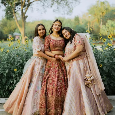 A bride stands happily with her two sisters, all in beautiful lehengas, in a sun-drenched garden.