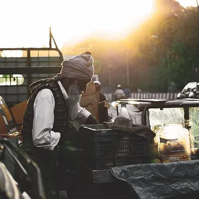 An elderly street vendor with a long white beard arranges his goods in the beautiful golden hour light. This candid street photograph captures a quiet moment of daily work in Bangalore.