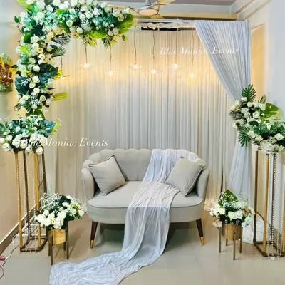 A breathtaking Roka setup featuring a lush floral arch of white roses and green leaves, with hanging lights creating a magical glow. The decor is complemented by floral stands and a classic sofa.