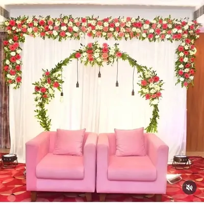 A beautiful stage backdrop for an engagement ceremony featuring a clean white curtain. The decor is highlighted by a lush floral arch of red and white flowers with hanging Edison bulbs, perfectly framing the two pink armchairs for the couple.