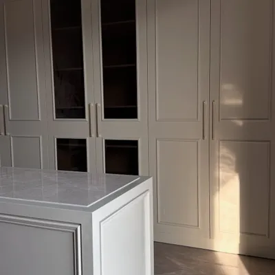A beautifully lit walk-in closet with off-white paneled wardrobes and a central island with a marble top. The natural light highlights the clean lines and elegant finish of our custom woodwork.