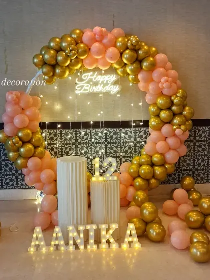 A beautiful setup for a 12th birthday, featuring a peach and gold balloon ring, a neon sign, fairy lights, and the name 'Aanika' in marquee letters.