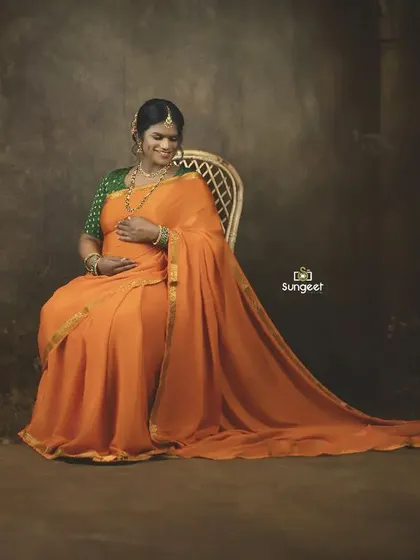 A serene, seated portrait of the mom-to-be in an orange saree. The dark, moody background makes her and the baby bump the central focus.