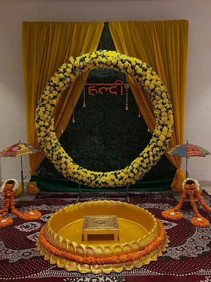 A beautiful haldi setup featuring a large circular arch of yellow flowers against a green backdrop. The lotus seating area is placed on a traditional carpet.