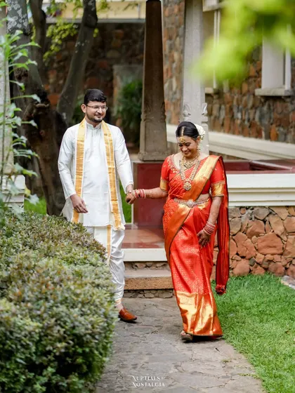 A newly married couple in traditional South Indian attire walks hand-in-hand through a beautiful garden courtyard after their wedding.