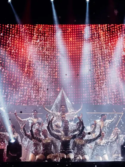 This shot captures the energy of a grand finale. The stage is alive with a glittering LED background, powerful spotlights, and dancers in a final, striking pose around the central pyramid structure.