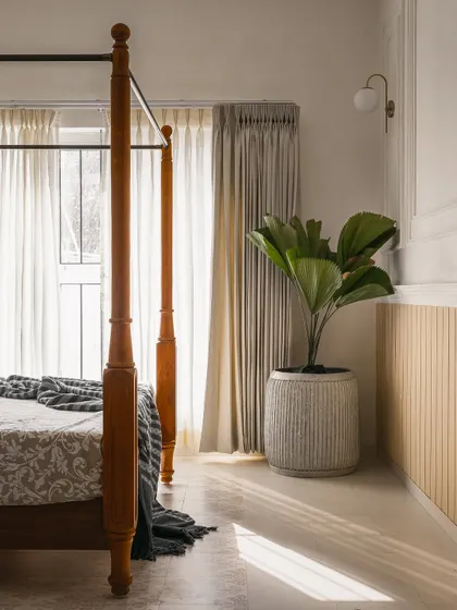 Natural light filters through sheer curtains, illuminating a corner of the bedroom. The space is kept simple with a fiddle-leaf fig plant and minimalist wall paneling to complement the traditional bed frame.