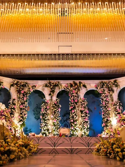 A full view of an elegant reception stage featuring floral-draped arches against a soft blue backdrop. The design, illuminated by the grand ballroom chandeliers, creates a feeling of classic romance.