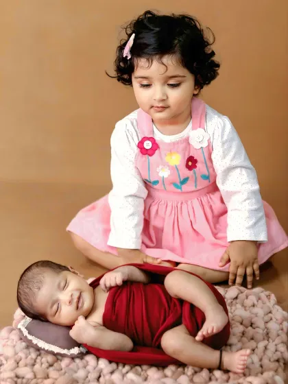 A touching moment between siblings, as a big sister in a pink dress looks lovingly at her sleeping newborn sibling.