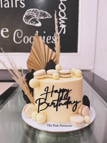 An elegant cream-colored birthday cake with modern black accents. This design features a bold "Happy Birthday" topper, white macarons, and dried floral elements for a contemporary look.