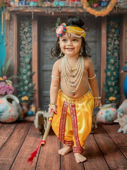 A happy toddler dressed as Krishna, standing confidently with his flute.