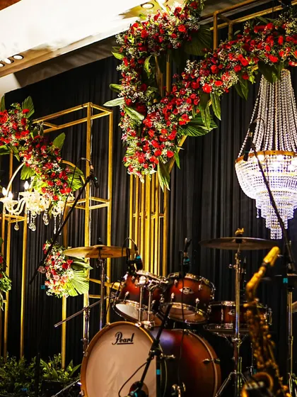 The stage is set for a memorable night of music. The drum kit is framed by golden arches, cascading red florals, and a sparkling crystal chandelier.