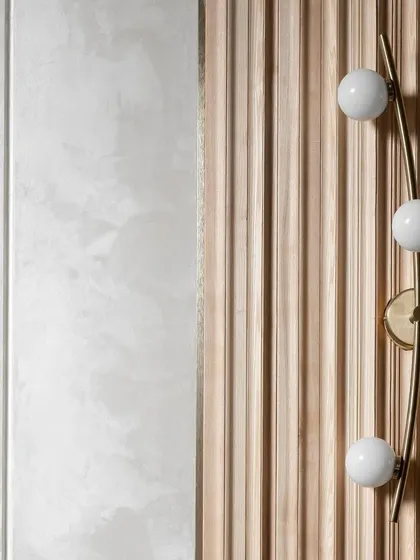 A study in texture and light. This composition features a modern brass wall sconce with globe lights set against a fluted wooden panel, which contrasts with the smooth, lime-plastered wall beside it.