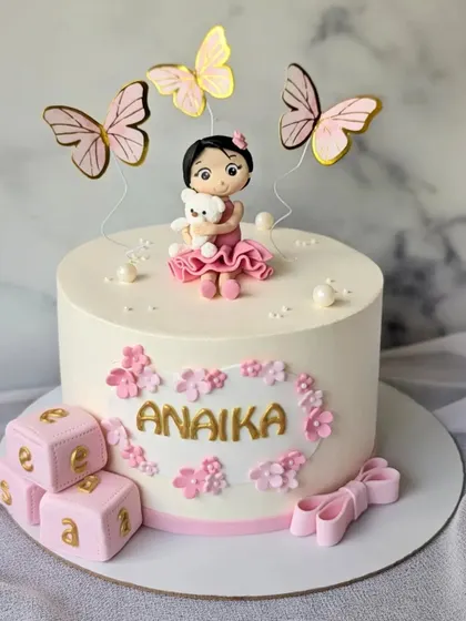 A beautiful cake for a baby girl's name reveal ceremony. The design is soft and feminine, with a custom figurine, pink blocks, and butterflies.