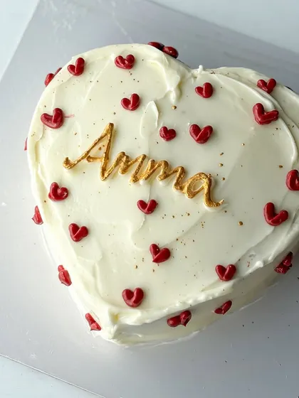 A simple and heartfelt cake for Amma. This heart-shaped cake is covered in white buttercream and decorated with tiny red hearts and a beautiful gold "Amma" script.