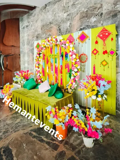 An indoor Mehendi or Haldi corner setup against a marble wall. The bright green and yellow backdrop is decorated with kites and floral arrangements for a festive look.