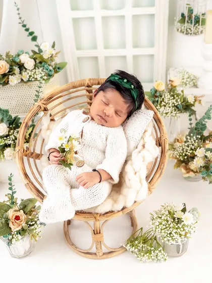 A beautiful overhead shot of a baby sleeping in a round wicker chair, surrounded by delicate flowers. The green headband adds a lovely pop of color.