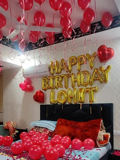 A classic red balloon birthday decoration for a partner, with "Happy Birthday" and the name "Lohit" in gold foil letters over the bed.