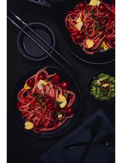 A dark, moody shot of two plates of red-hued noodles, styled with cherry tomatoes and edible flowers. The use of black tableware and chopsticks creates a modern, Asian-inspired culinary scene.