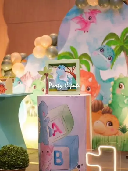 A close-up of the dessert table area for Avyaan's dino party. The setup includes a custom backdrop with cartoon dinosaurs and themed pedestals for the cake and treats.
