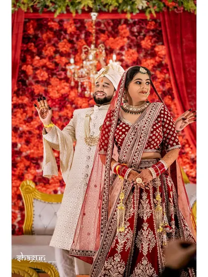 A happy, celebratory pose from the bride and groom on their wedding stage, against a vibrant red floral backdrop.
