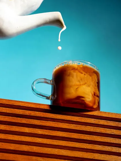 An artistic shot of milk being poured into a cup of coffee against a bright blue sky. The contrast between the warm coffee and the cool background creates a visually striking and refreshing image.