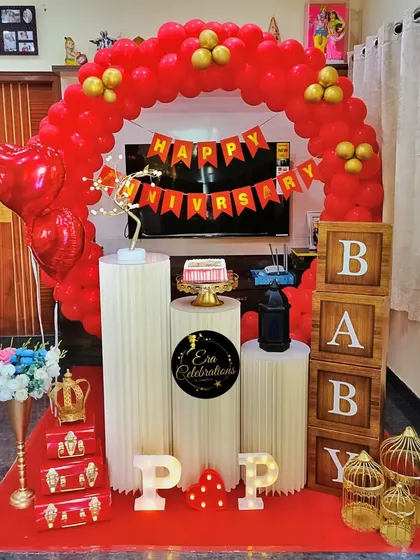 A vibrant red and gold anniversary decoration for a home celebration. The setup includes a red balloon arch, heart-shaped balloons, and decorative props.