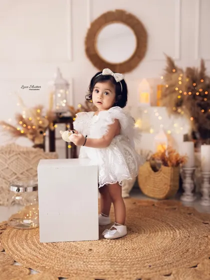 An elegant and modern first birthday portrait. This little girl in a beautiful white lace dress stands in a chic, boho-inspired setting with warm lights.