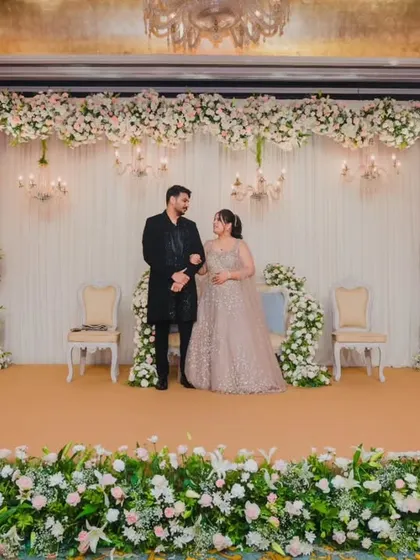 A wide shot of the couple on their beautifully decorated reception stage, showcasing the grand floral arrangements and elegant chandeliers.