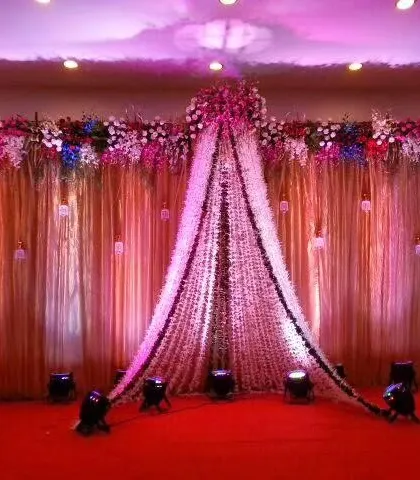 An elegant reception stage with a backdrop of flowing drapes and a canopy made of flower strings, beautifully illuminated by spotlights.