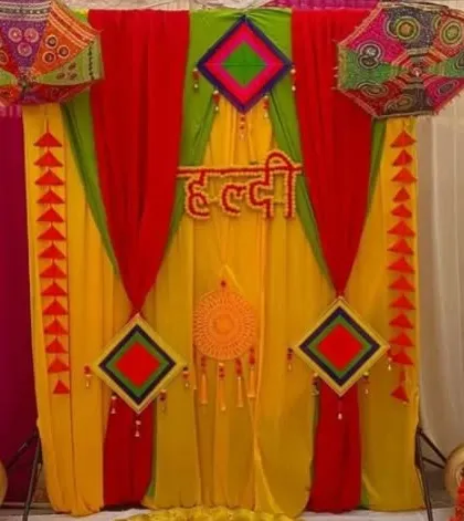A Haldi backdrop with red, yellow, and green drapes, decorated with colorful kites and a central dreamcatcher.