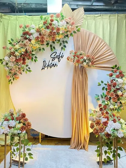 A stylish engagement backdrop with a large white circular panel and earthy-toned floral arrangements. A touch of gold fabric adds a bit of shimmer, and the couple's names can be added for a personalized touch.