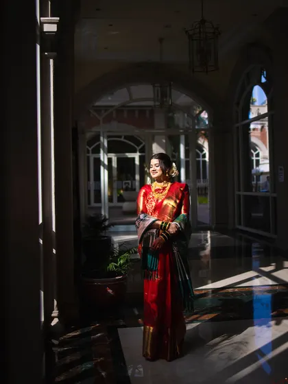 An artistic portrait of a bride in a red saree, beautifully lit by streams of sunlight in an elegant corridor.