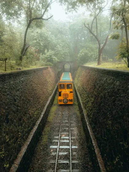 The toy train at Sanjay Gandhi National Park emerges from a tunnel, framed by symmetrical stone walls. The leading lines of the track draw the eye into the lush, green landscape.